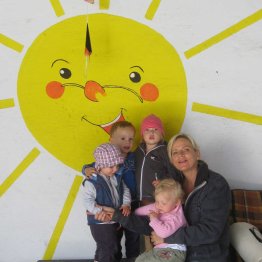 Das Bild zeigt die Pflegekraft von der Tagespflege Sonnenschein zusammen mit 4 Kindern vor einer Wand, auf der eine Sonne gemalt ist