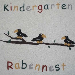 Das Bild zeigt das Logo der Kindertagesstätte Rabennest auf der Fassade des Kita-Gebäudes. Das Logo zeigt 3 Vögel sitzend auf einem Ast.